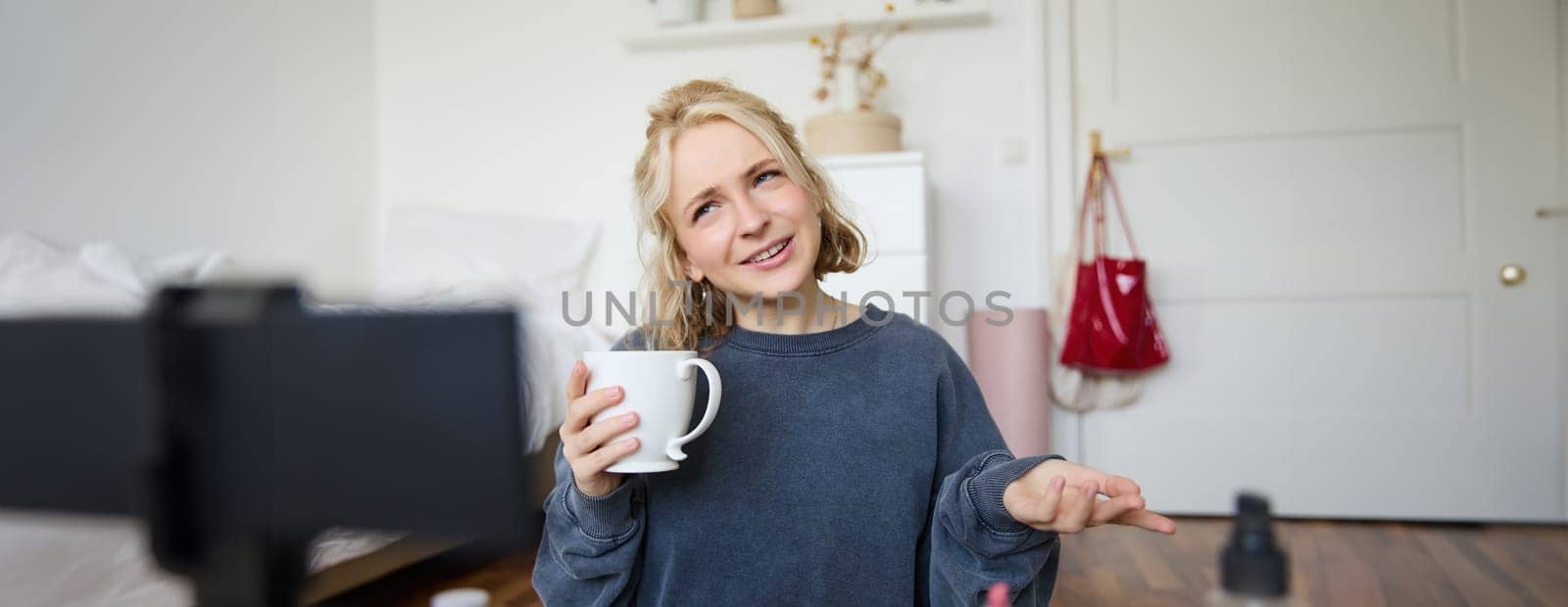 Portrait of blond young lifestyle blogger, woman records video of her talking about life and beauty, sits in front of camera, holds cup, drinks tea, does blog content for social media account by Benzoix