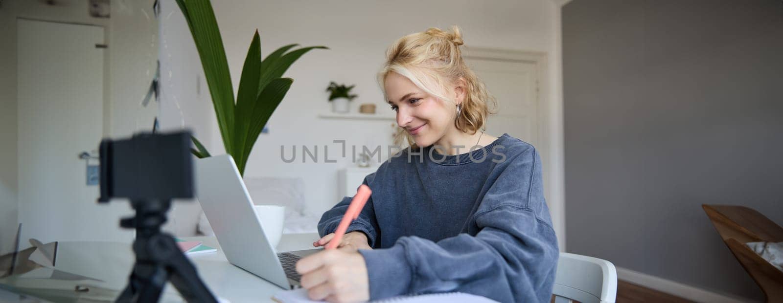 Portrait of smiling blond woman writing in notebook, making notes, recording content for social medial on digital camera, looking at laptop, working or studying.