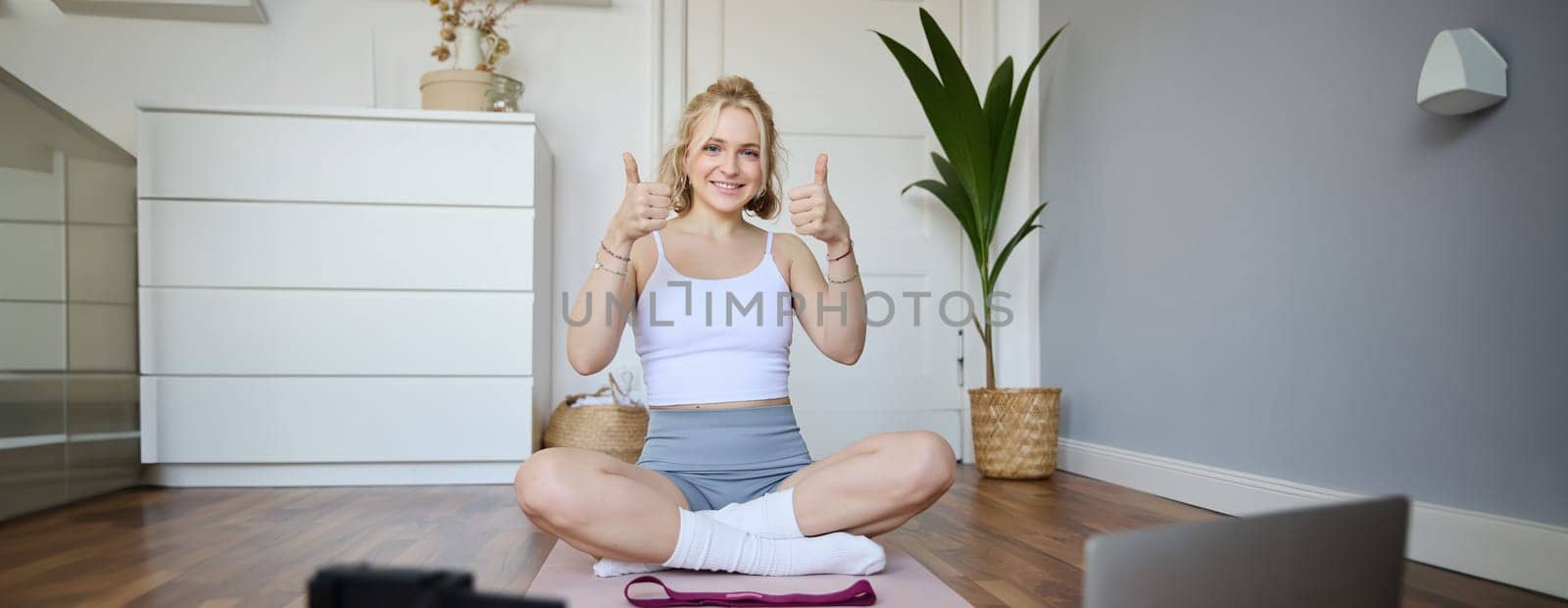 Healthy and fit fitness blogger, recording vlog for wellbeing social media account, sitting on yoga mat and showing thumbs up, using digital camera to create content, following script of laptop.