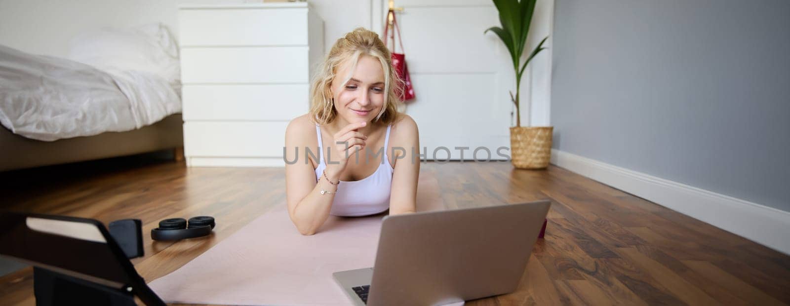 Portrait of young fitness woman, doing workout at home, looking at her gym instructor on laptop screen, doing online training session by Benzoix