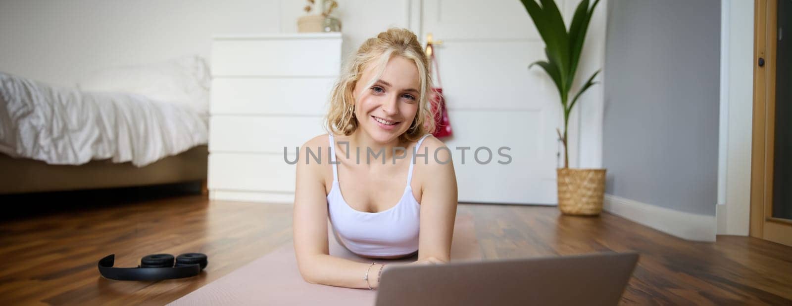 Portrait of cute young fitness woman, doing workout at home, lying on rubber yoga mat, watching exercises online on laptop.