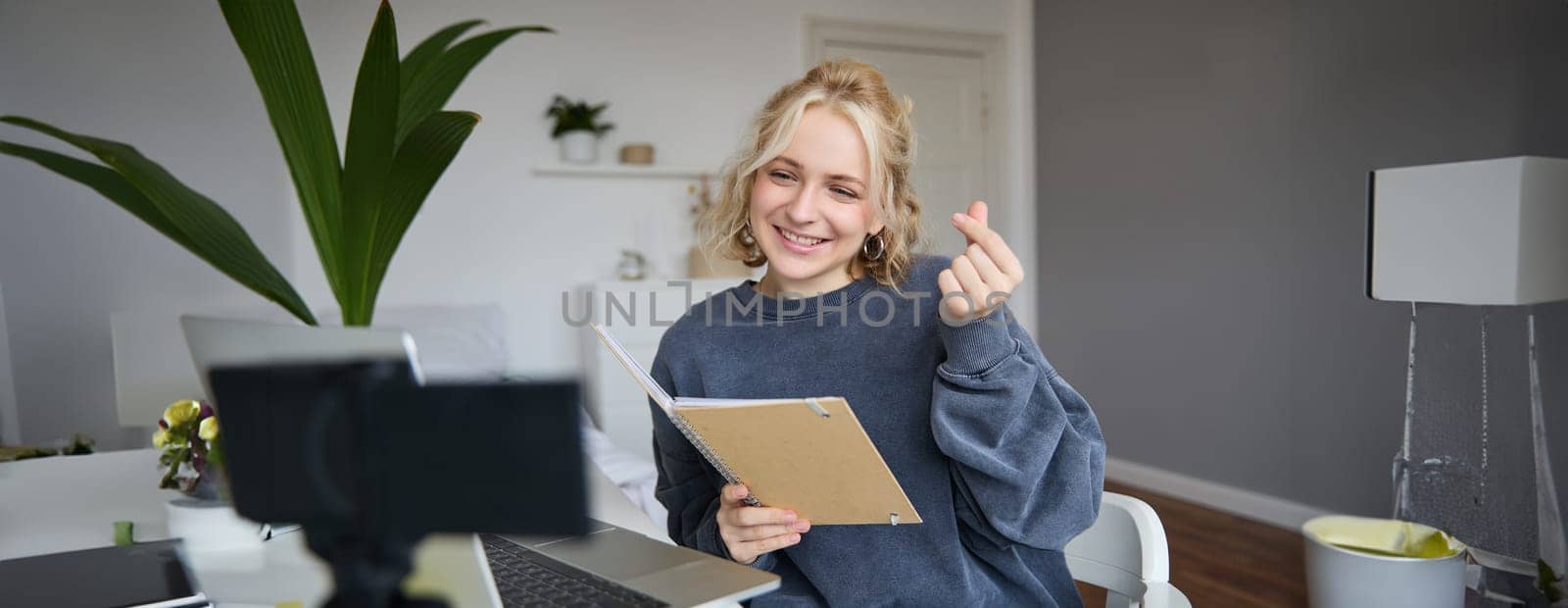 Cute young woman in her room, holds notebook, sits with laptop, looks at digital camera, records vlog or online tutorial.