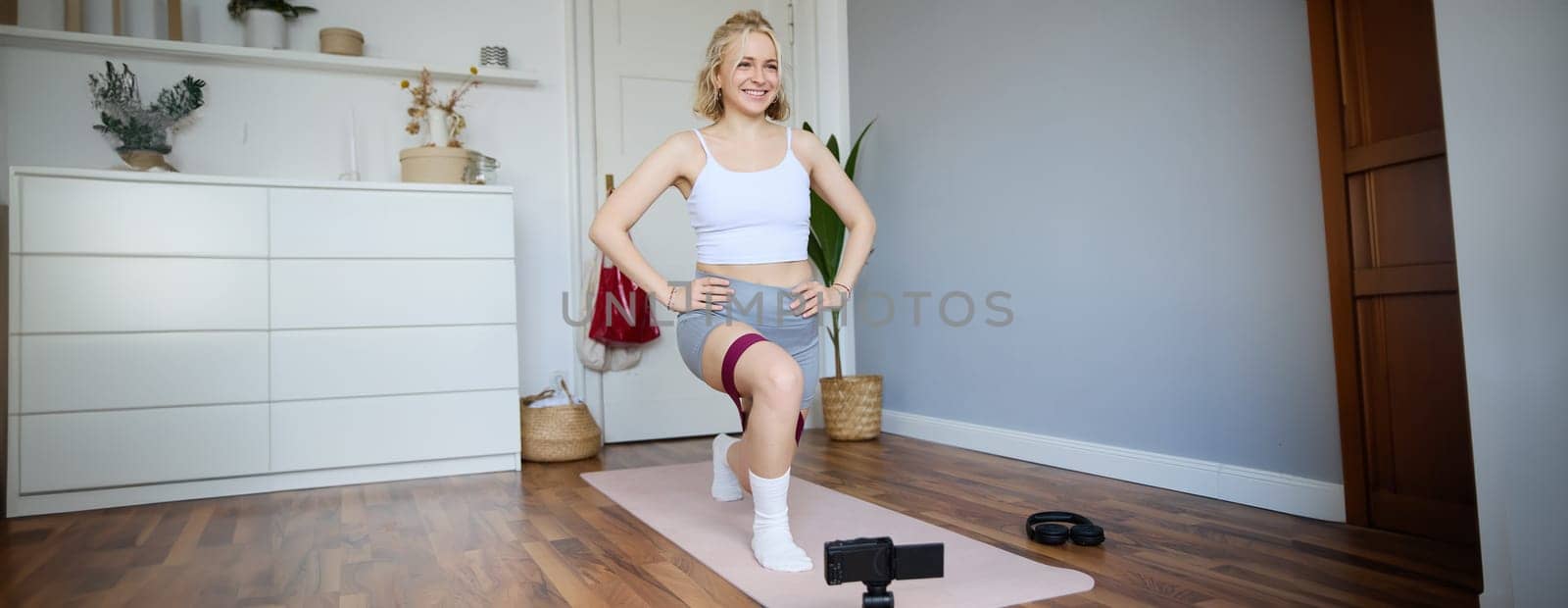 Portrait of young athletic woman, vlogger making a video about home workout, fitness instructor shooting how to do leg exercises, using elastic resistance band, using yoga mat.