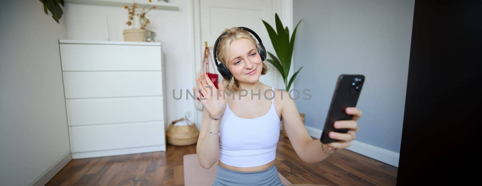 Portrait of young woman with smartphone and headphones, waving hand at mobile phone camera, live streaming during workout. Wellbeing and sport concept