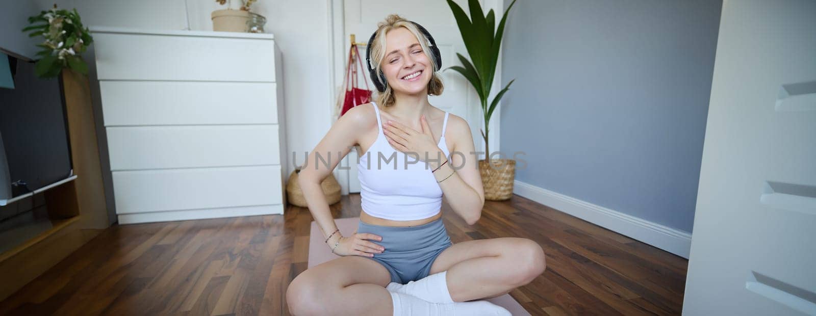 Portrait of young fitness woman, sporty female model in her room, sits on yoga mat, listens to music during workout, does exercises.