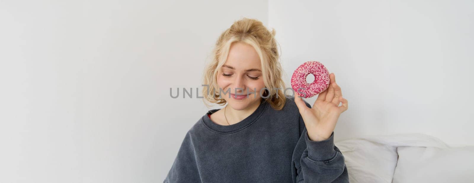 Portrait of cute blond girl holding pink doughnut with sprinkles on top, showing her favourite food.