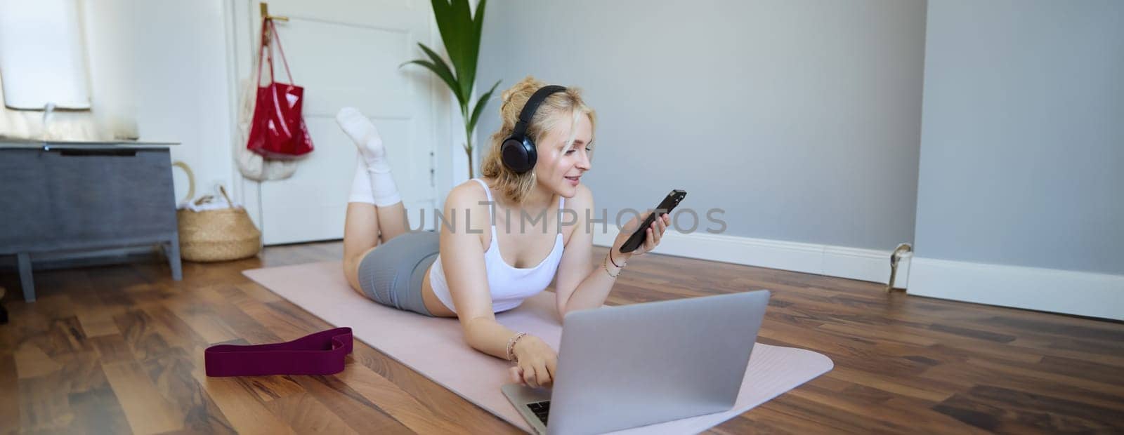 Portrait of young fitness woman, choosing workout on laptop, wearing headphones and using mobile phone exercise app. Sport and lifestyle concept