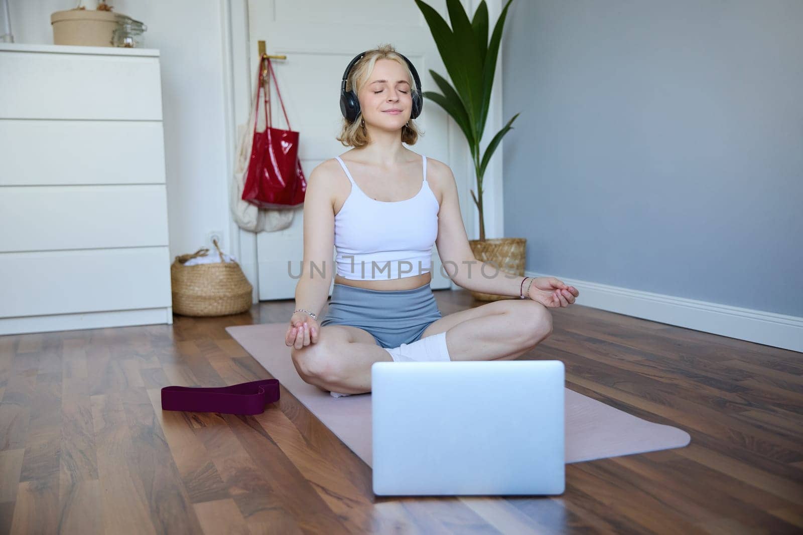 Portrait of concentrated woman in headphones, meditating, listening to calm music and practice yoga, follow instructions on laptop, sitting on rubber mat by Benzoix