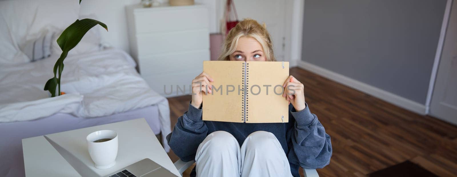 Portrait of cute blond woman, sits in front of digital camera and laptop in her room, covers face with notebook journal, records video of herself for lifestyle vlog.