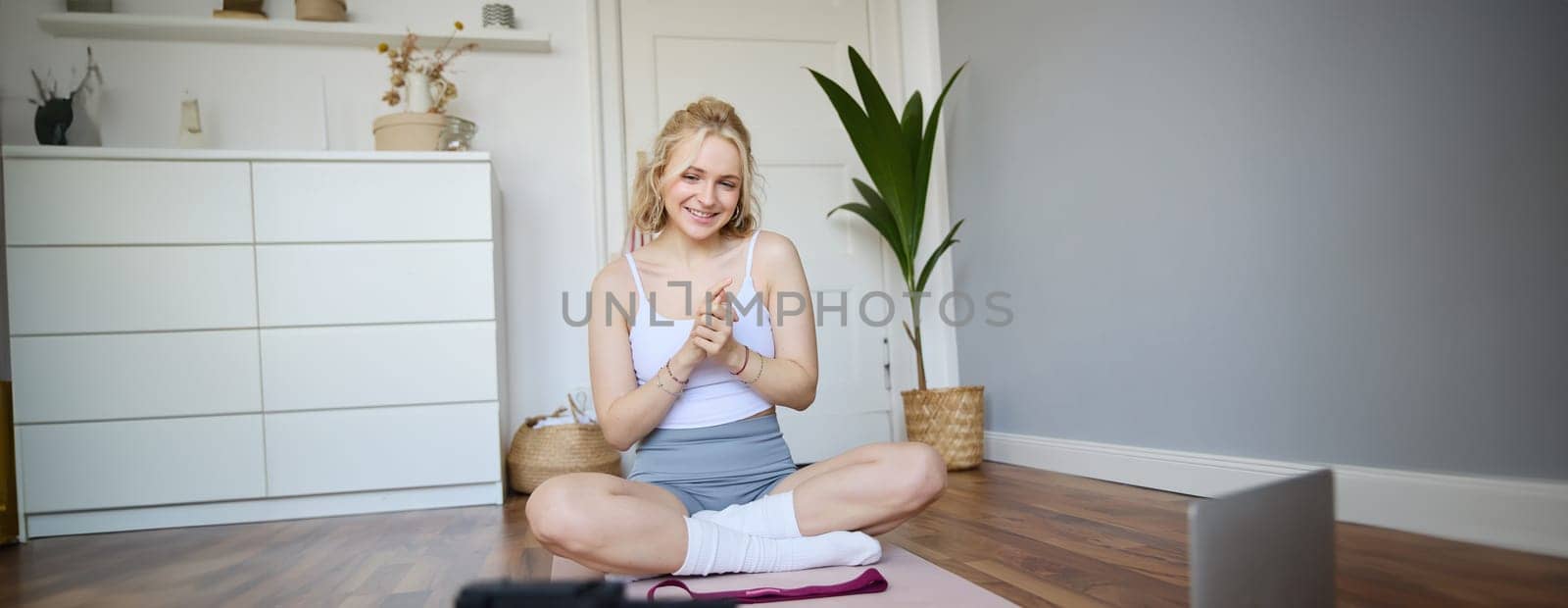Portrait of young charismatic fitness trainer, girl blogger records video on digital camera, talks about health and workout, doing exercises on rubber mat in a room at home by Benzoix