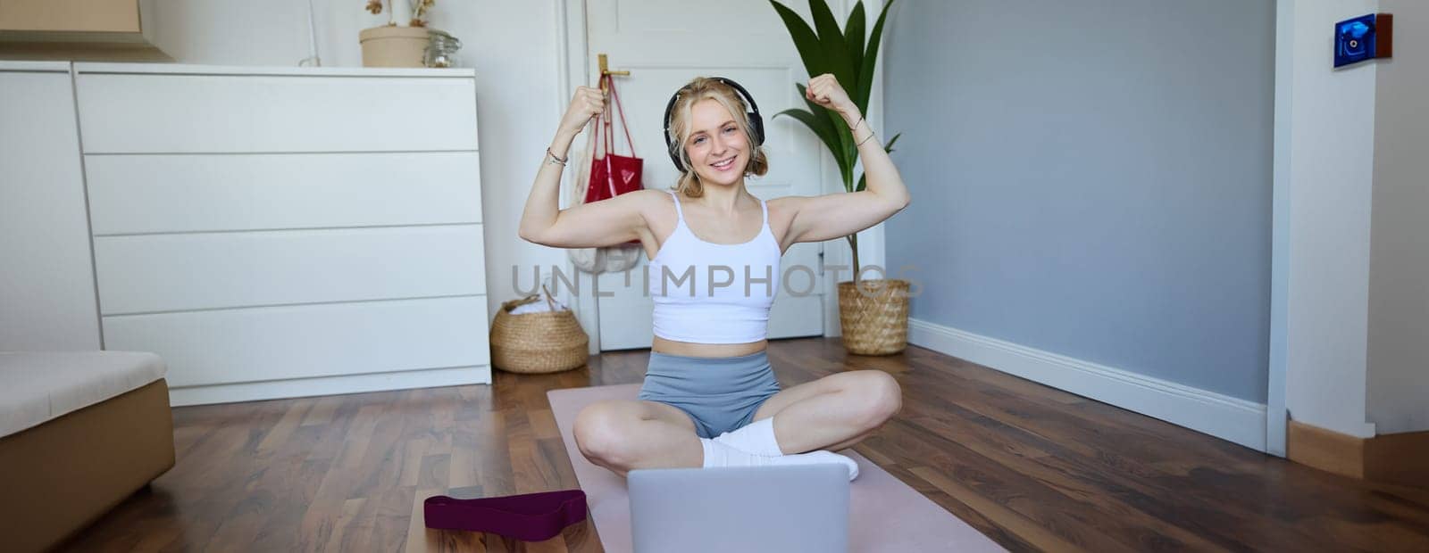 Portrait of fit and healthy, sporty woman showing her muscles on camera, wearing wireless headphones, using laptop for home workout training by Benzoix