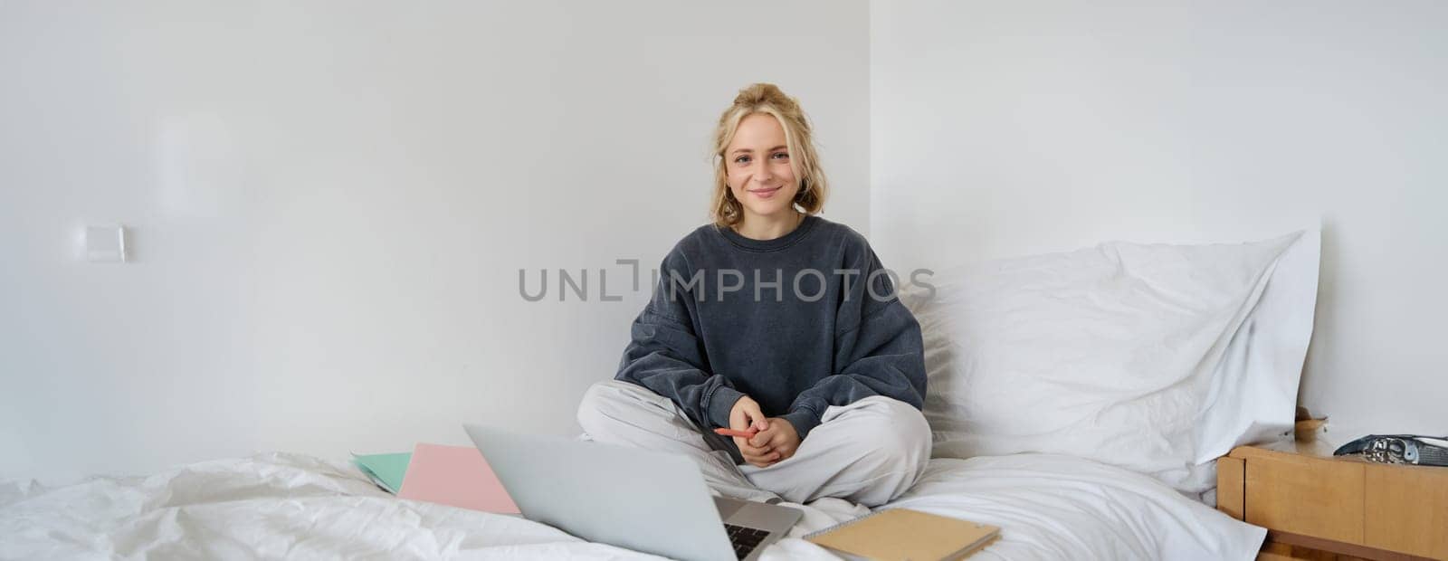 Portrait of happy blond woman, freelancer working from home, sitting on bed with laptop and notebooks. Student doing homework in bedroom, connects to online class via video chat.