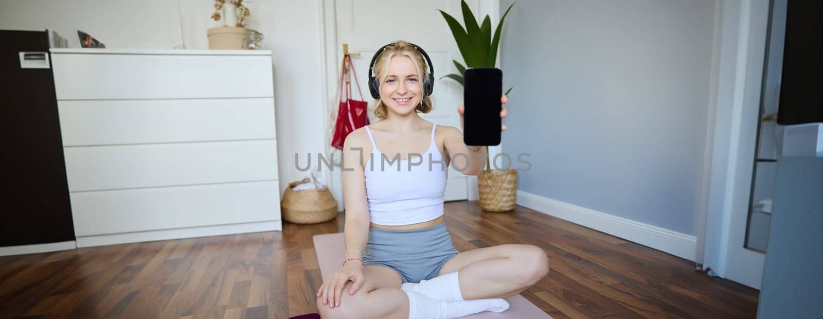 Portrait of beautiful young fitness woman, wearing headphones, showing workout app on smartphone, demonstrating mobile phone screen and smiling, sitting on yoga mat by Benzoix