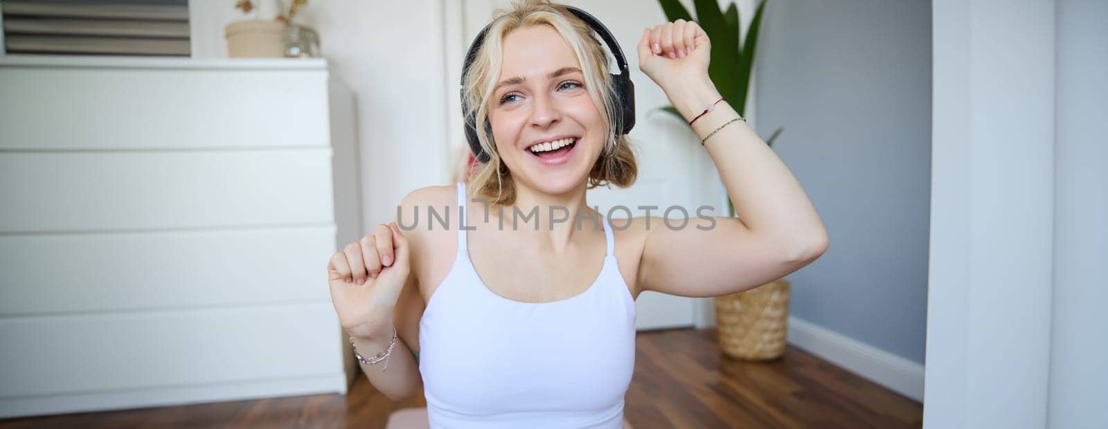 Portrait of charismatic, beautiful young woman dancing and listening to music in wireless headphones, singing along favourite song in her room by Benzoix
