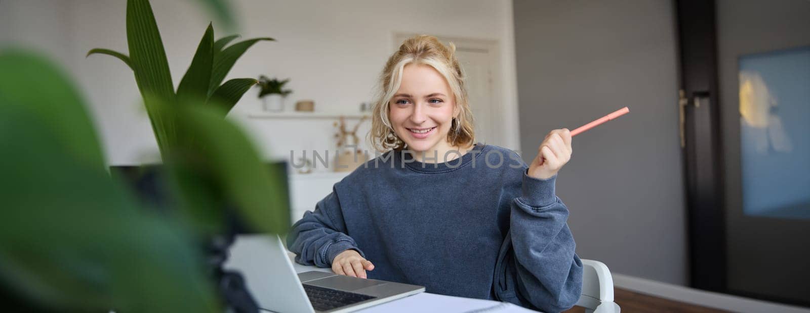 Portrait of young charismatic woman, smiling, holding pen, looking at laptop, video chats, doing online course, answering a question, studying with tutor.