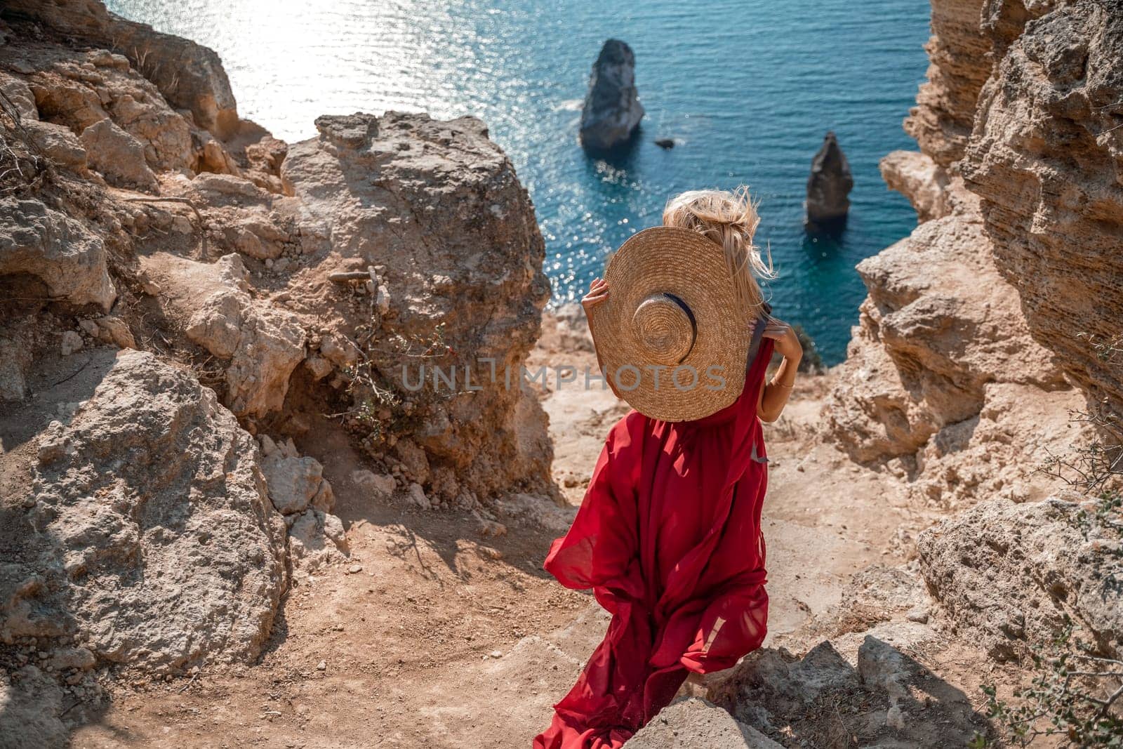 From behind, a woman is seen in a red flying dress fluttering in the wind. In a straw hat, walking down the stairs against the background of the sea with rocks and boats. The concept of romance, travel and tourism. by Matiunina
