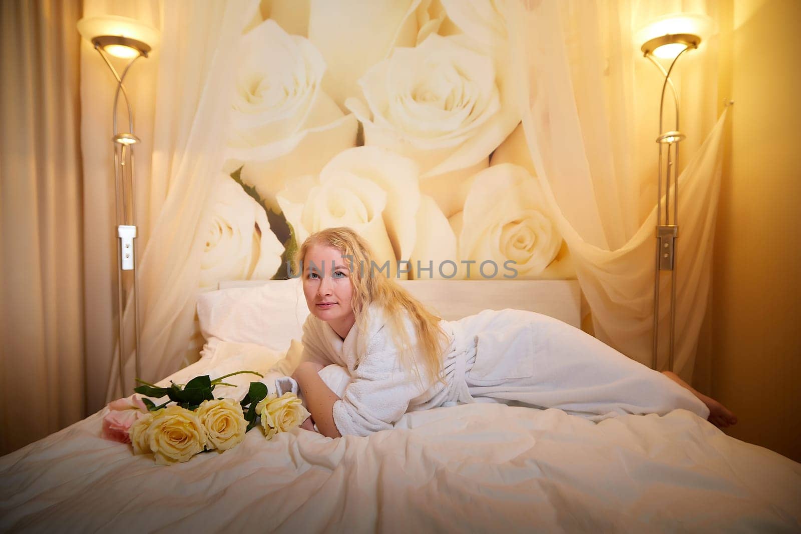 A blonde girl in a white terry bathrobe in a hotel room with flowers. Wedding travel, Weekend and vacation, body care by keleny