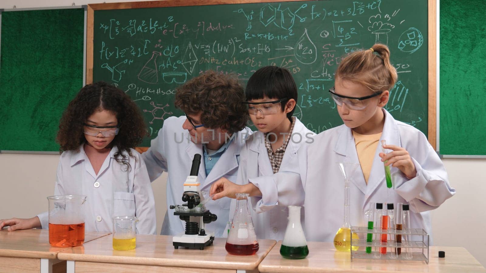Smart caucasian teacher looking under microscope while diverse children doing experiment at STEM science lesson. Professional happy scientist inspect biochemical example by using microscope. Pedagogy.