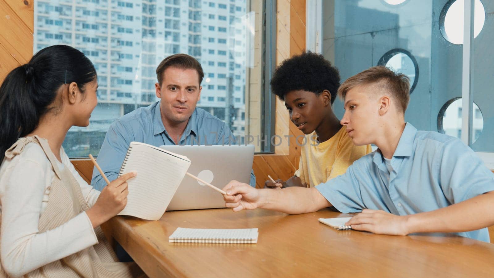 Smart girl ask teacher a question while show idea while diverse student working classwork. Group of multicultural children pointing at notebook while happy boy sharing idea at classroom. Edification.