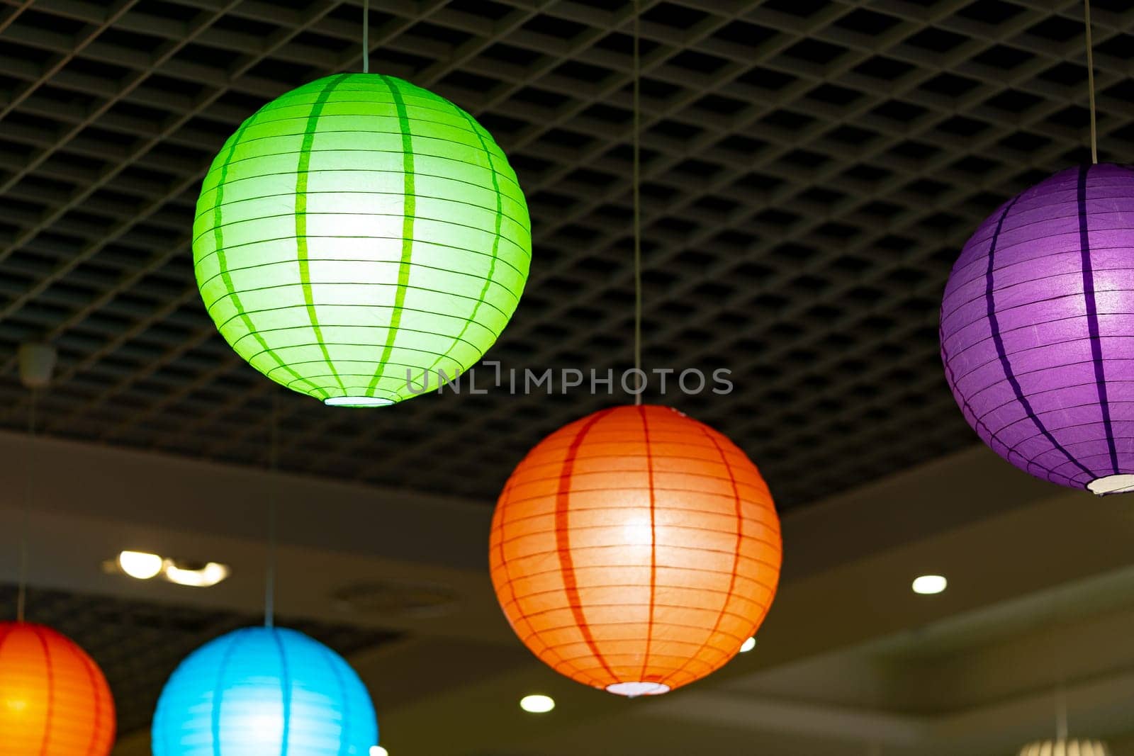 A collection of colorful paper lanterns hangs from the ceiling, gently illuminated from within. The lanterns, in hues of green, purple, orange, and blue, add a festive atmosphere to the indoor space, suggesting a celebration or cultural event.