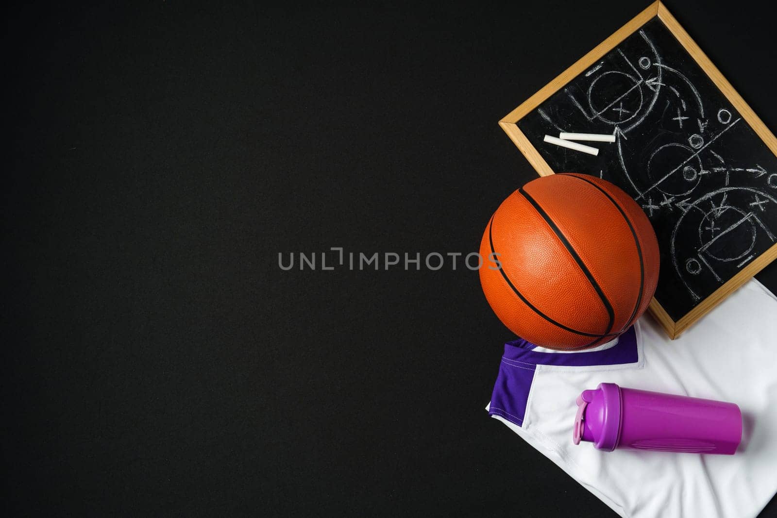 A basketball, along with a strategic play diagrammed on a chalkboard, rests near a folded jersey and a sports bottle. The equipment is arranged on a dark surface, suggesting preparation for a game or a coaching session in progress.