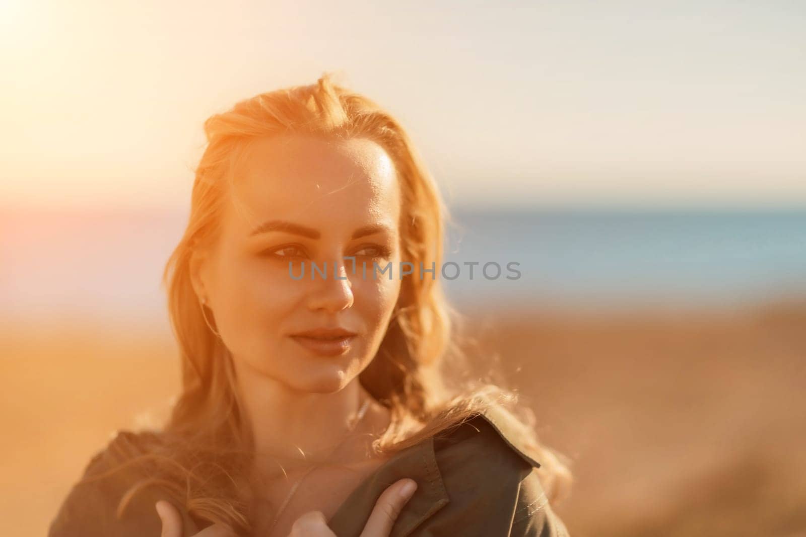 Portrait blonde sea cape. A calm young blonde in an unbuttoned khaki raincoat stands on the seashore, under the raincoat there is a black skirt and top by Matiunina