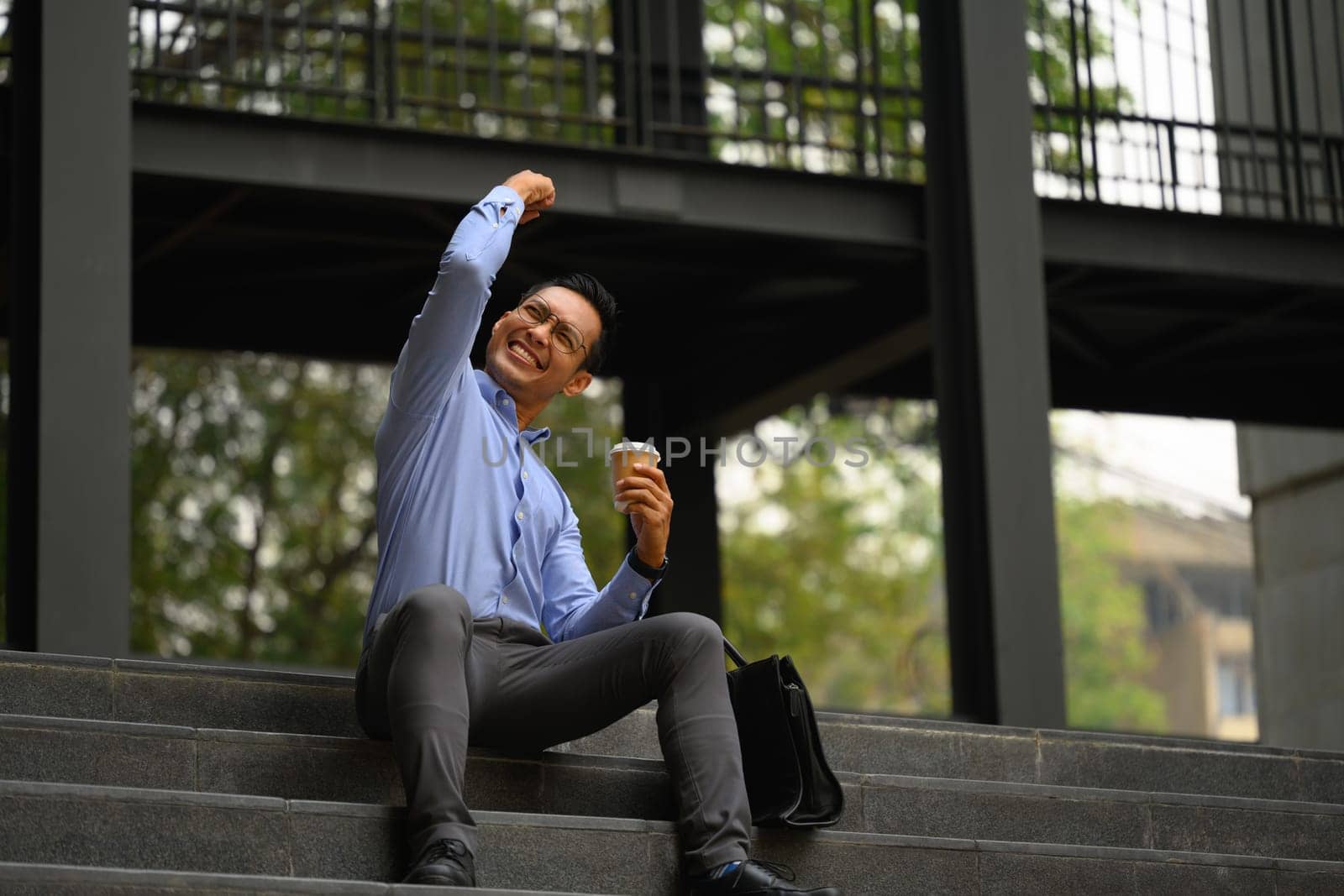 Excited young businessman celebrating success sitting outside office by prathanchorruangsak