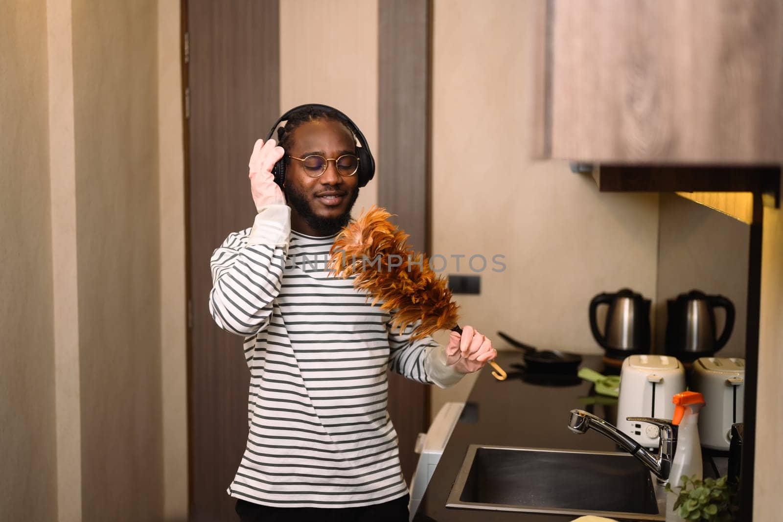Happy African man listening to music in headphones and singing while doing housework.