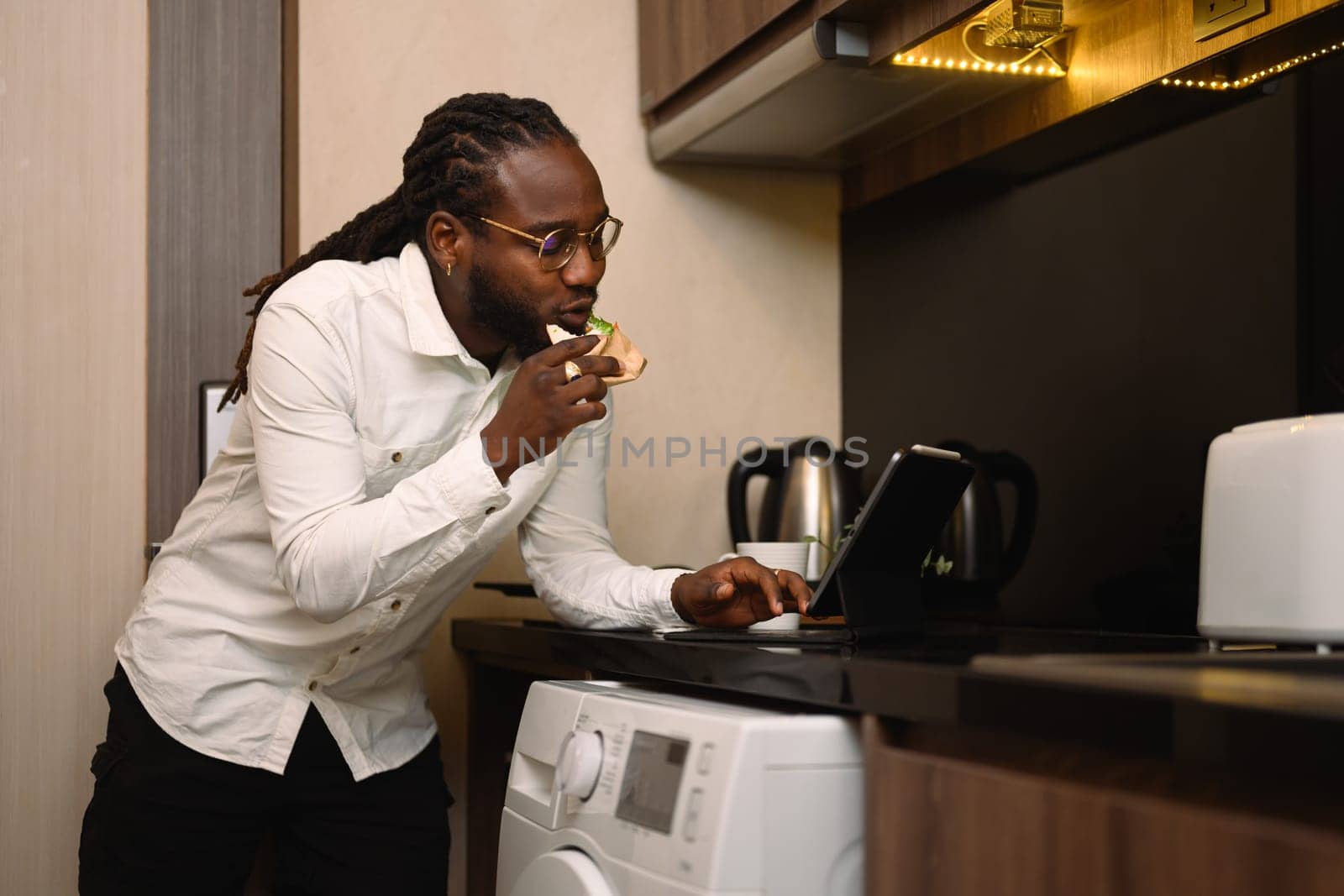 Portrait of African male in eyeglasses having breakfast in kitchen and using digital tablet.