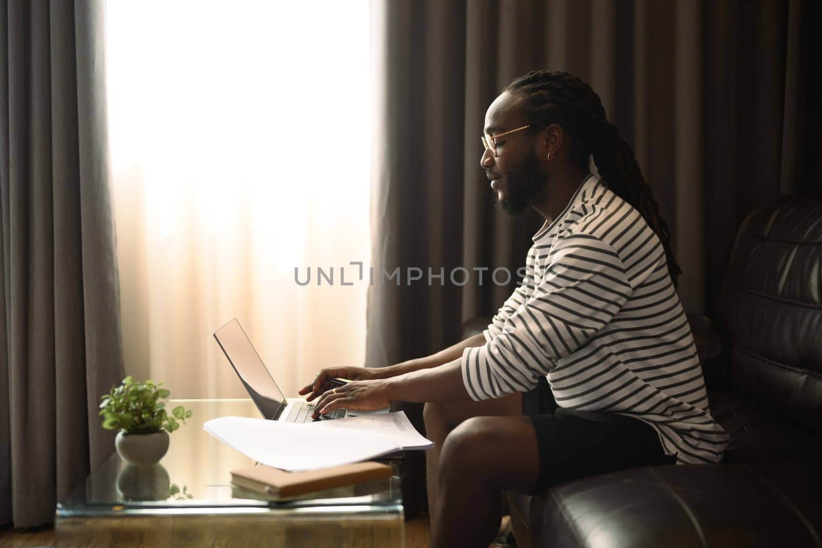 Young African man freelancer sitting on couch and working with laptop in living room.