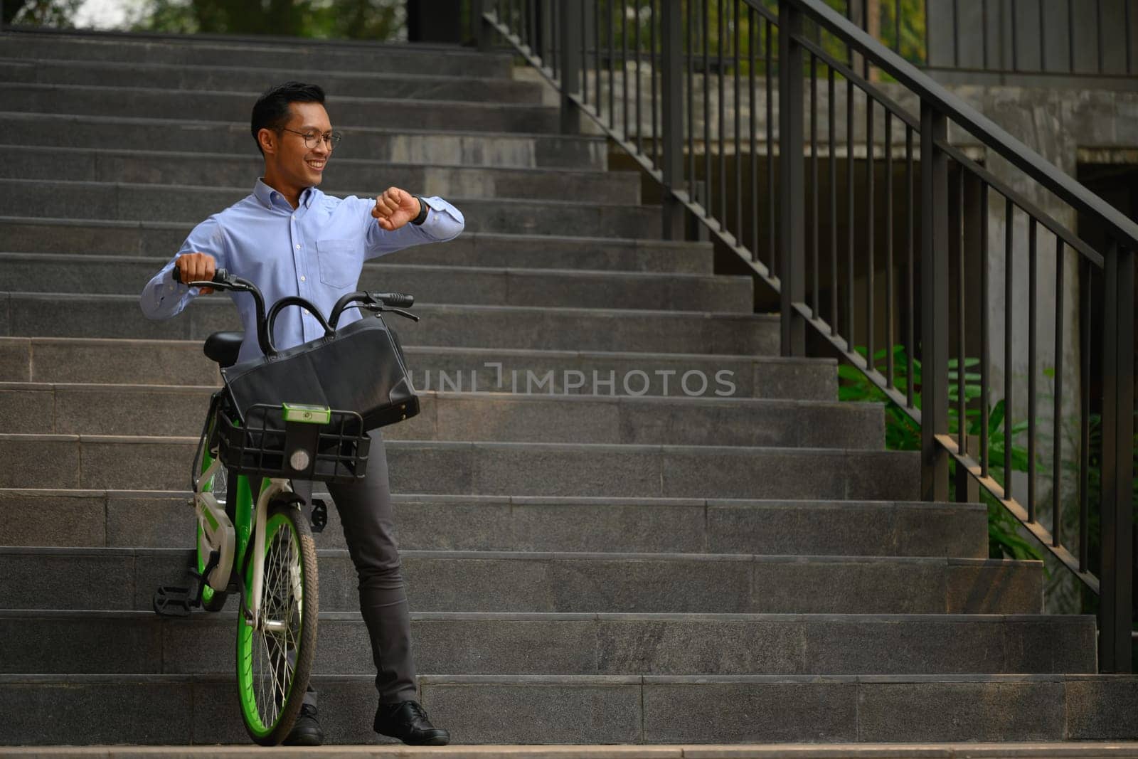 Smiling businessman with his bicycle in front of the building and checking time on wristwatch.
