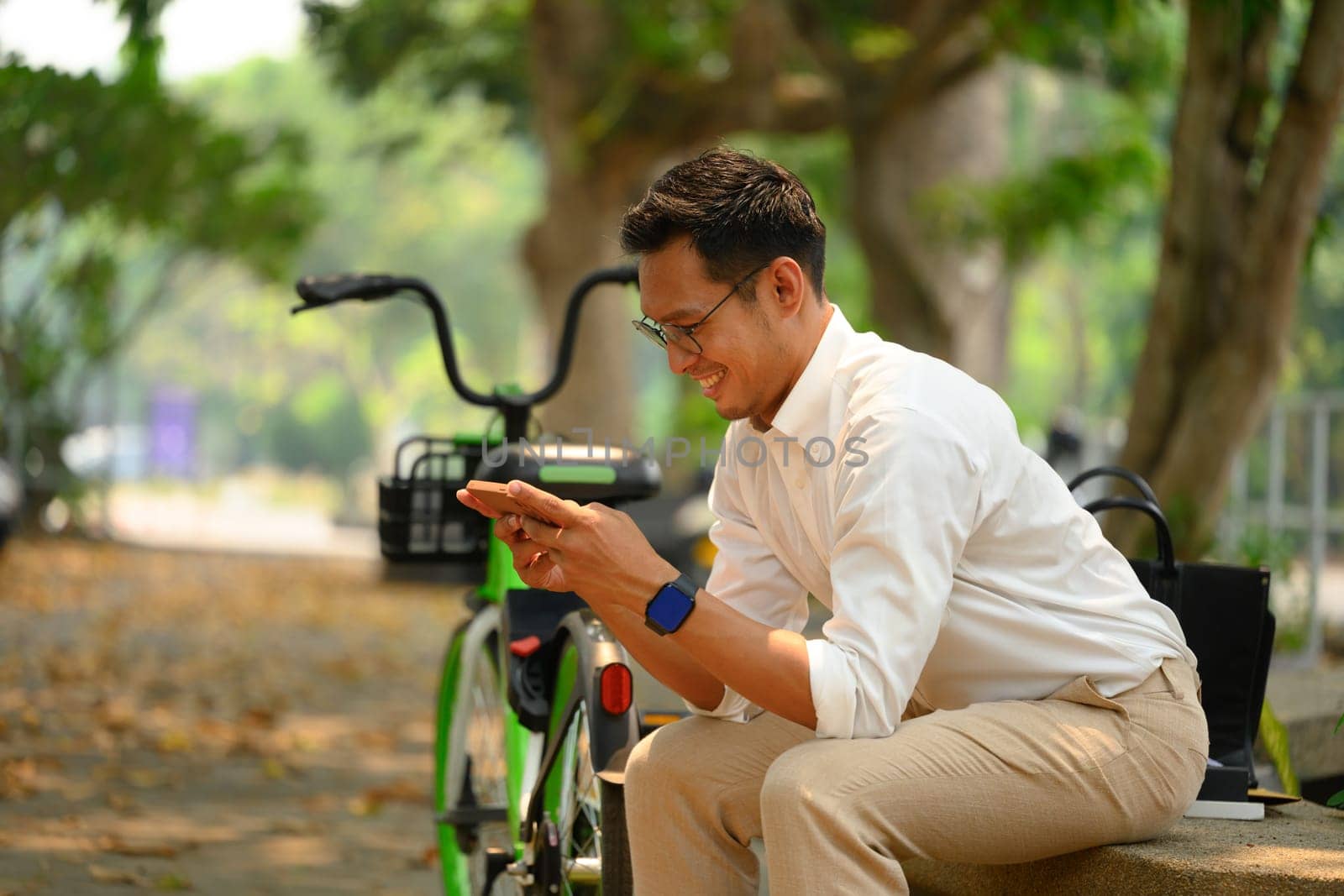 Happy young businessman sitting on bench near his bicycle using mobile phone by prathanchorruangsak