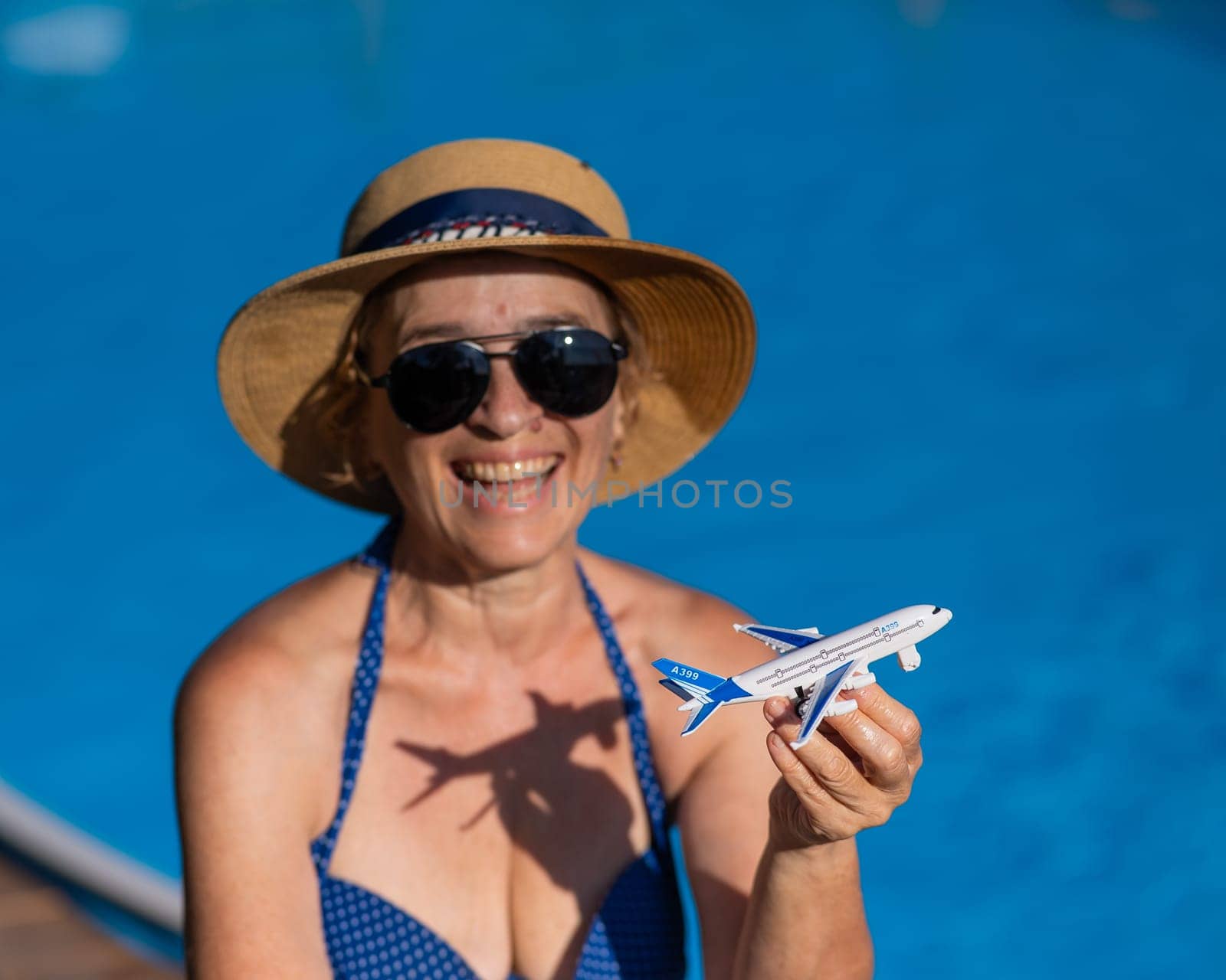 An elderly woman in a hat and sunglasses holds a model airplane while sitting by the pool. Retirement vacation concept