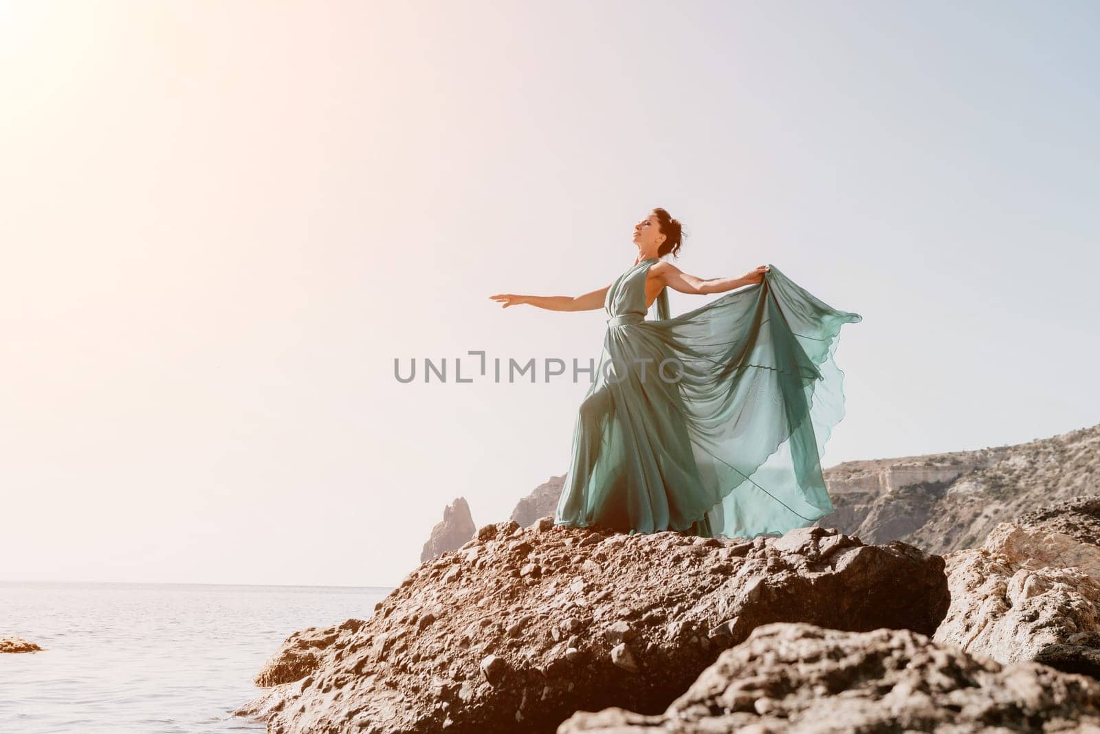 Side view a Young beautiful sensual woman in a red long dress posing on a rock high above the sea during sunrise. Girl on the nature on blue sky background. Fashion photo.