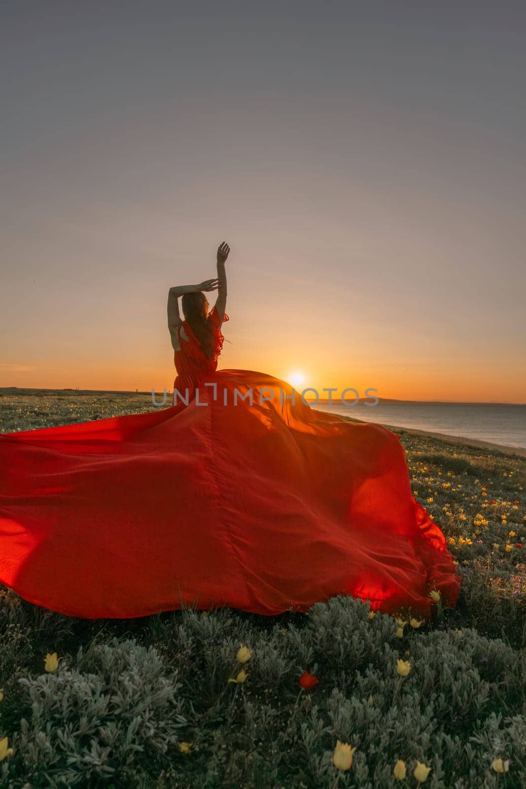 A woman in a red dress is standing in a field with the sun setting behind her. She is reaching up with her arms outstretched, as if she is trying to catch the sun. The scene is serene and peaceful