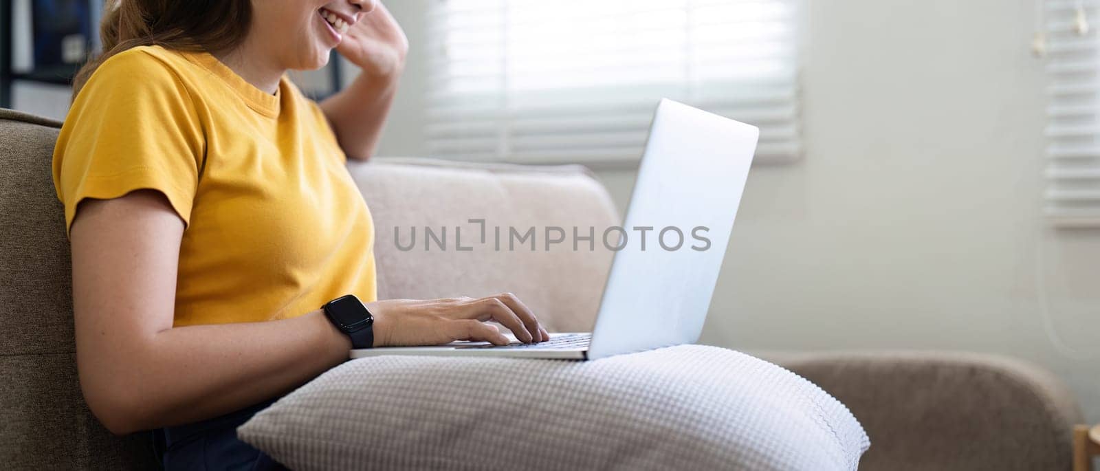 Young woman sitting on the couch and working on project, watching movie on laptop rest and happy chatting with friend in social network at home by nateemee