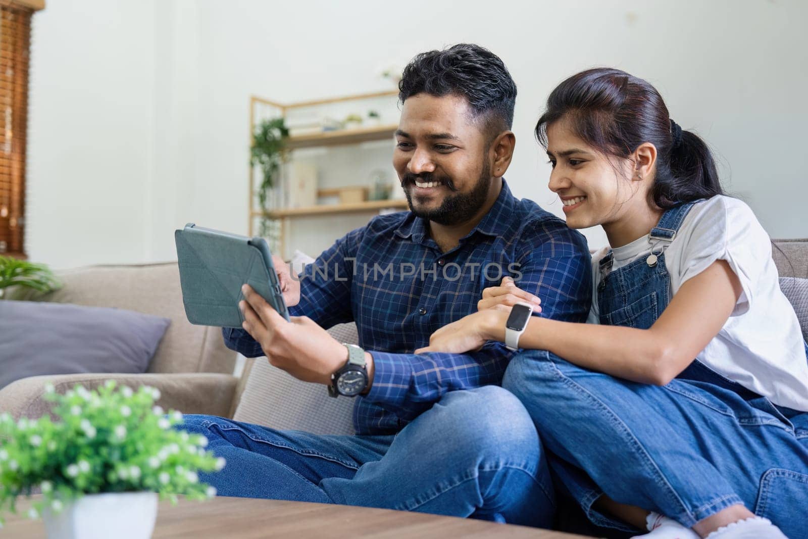 Indian married couple watching tablet together and sitting on sofa at home, Happy Indian husband and wife using tablet online services. Technology life style concept.