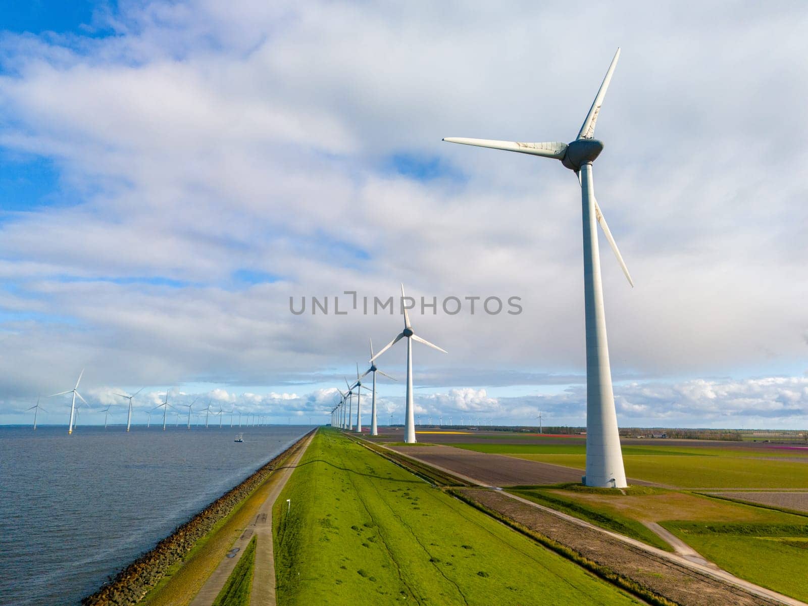 A picturesque scene of sleek wind turbines standing tall in a row next to a tranquil body of water, harnessing renewable energy on a bright Spring day by fokkebok