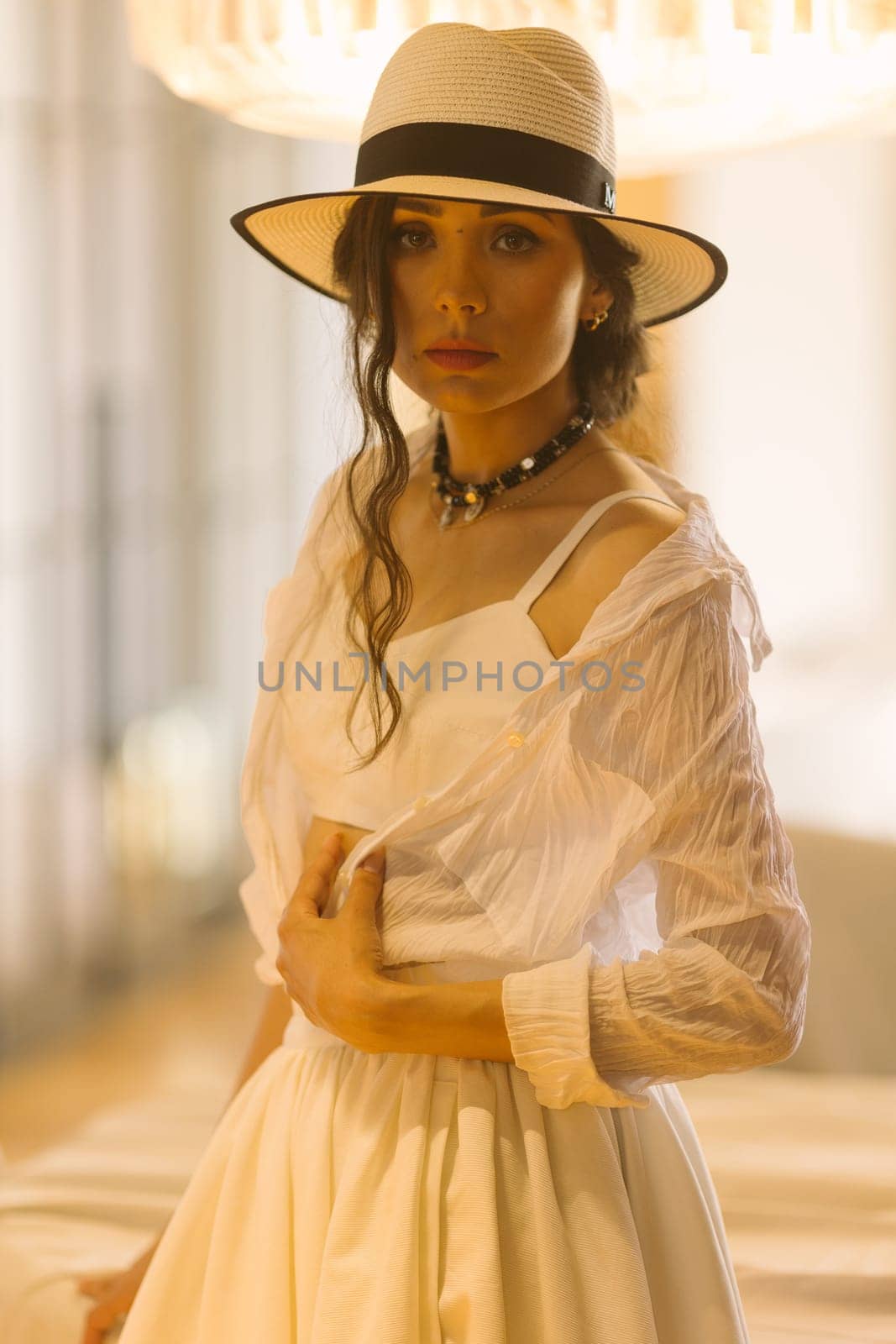 Elegant fashionable woman wearing summer white dress, straw hat, posing in stylish boho interior. Copy, empty space for text