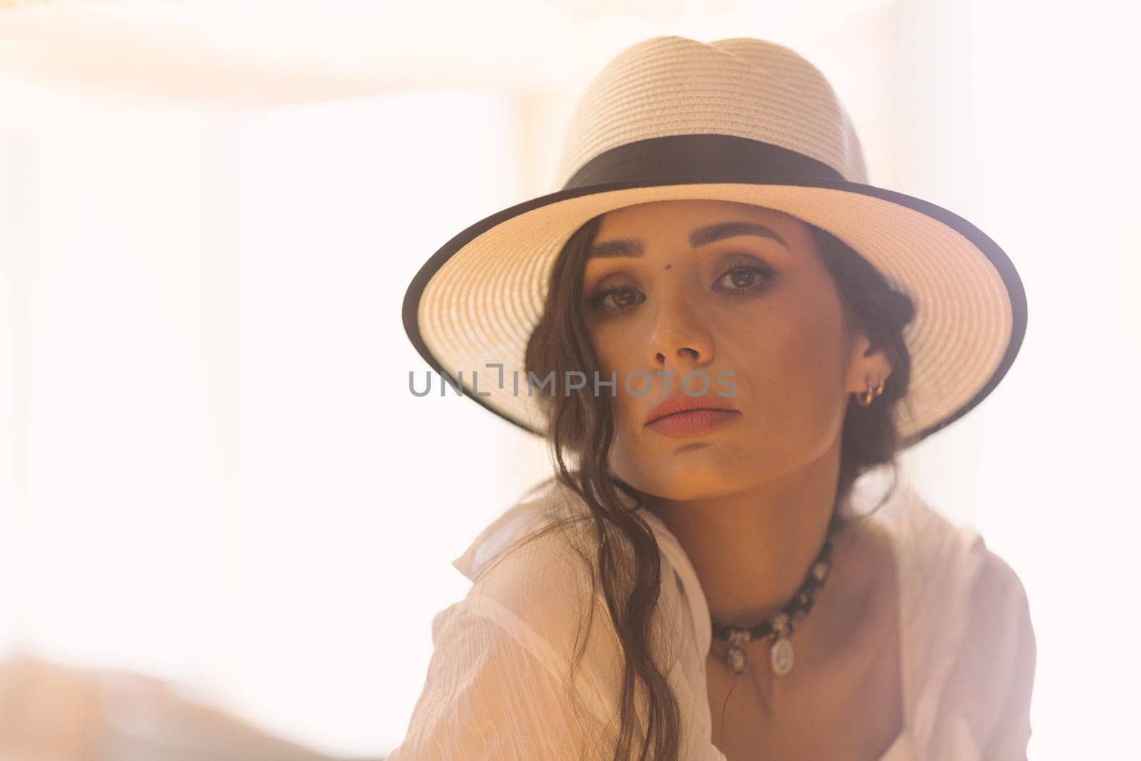 Elegant fashionable woman wearing summer white dress, straw hat, posing in stylish boho interior. Copy, empty space for text
