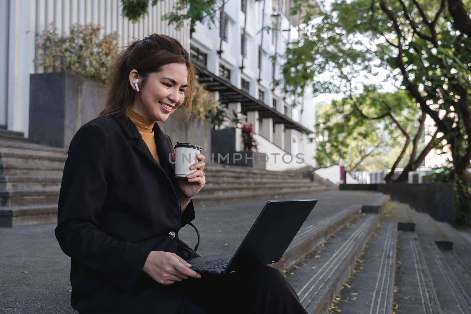 A woman in a black suit is sitting on a step with a laptop in front of her.