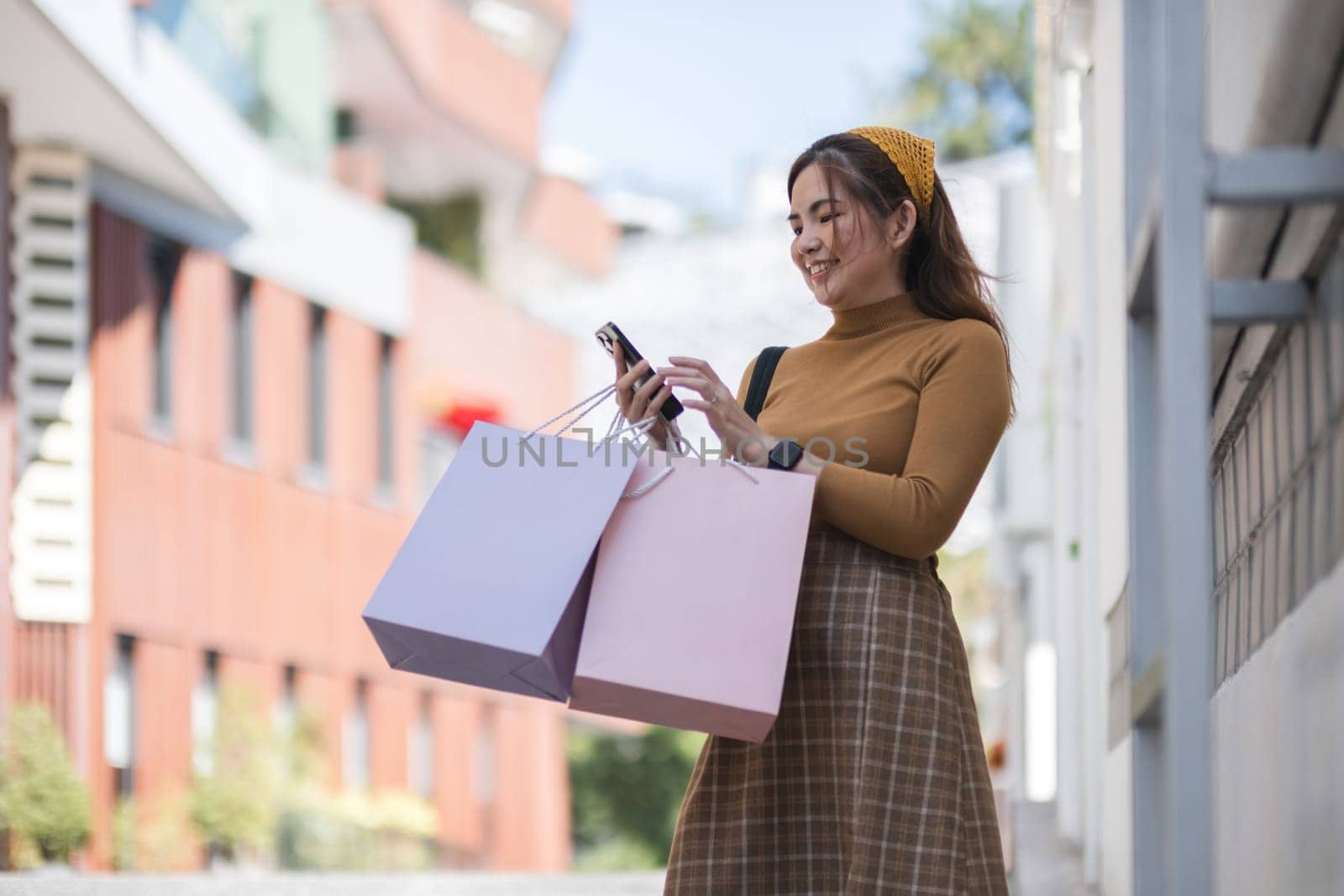 A woman is holding two pink shopping bags and looking at her cell phone by wichayada