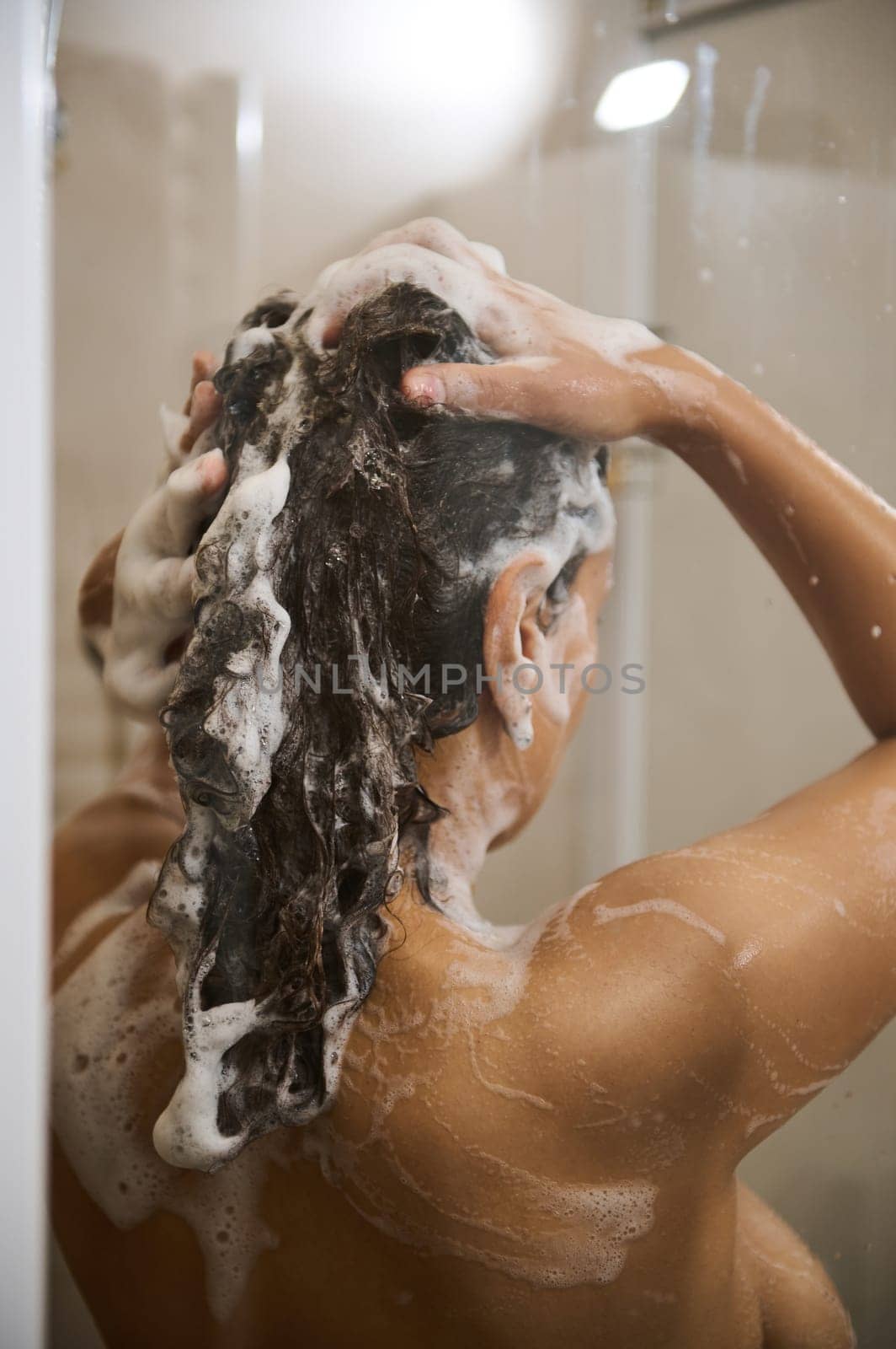 Rear view of pretty woman shampooing hair, showering with moisturizing foam for skin care and body freshness in a douche cabin. Close-up by artgf