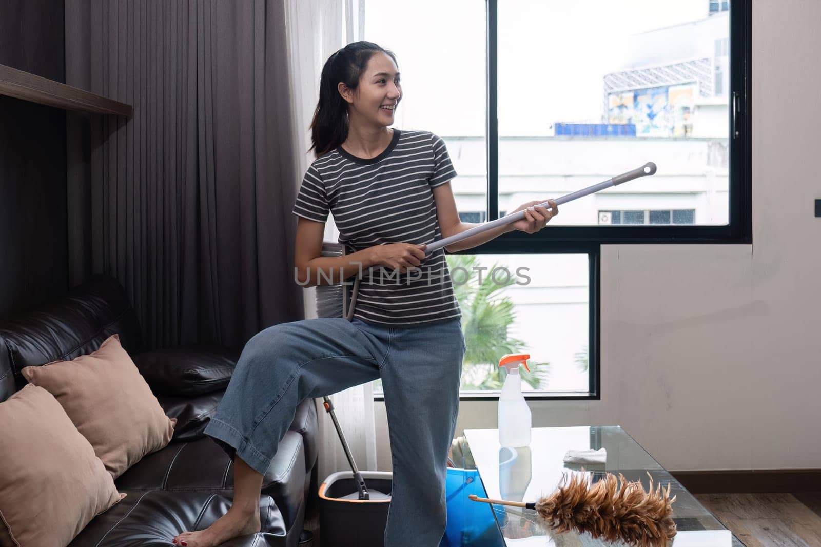 Cheerful and happy young woman singing and dancing while cleaning the living room, holding house cleaning equipment and wearing headphones, singing happily..