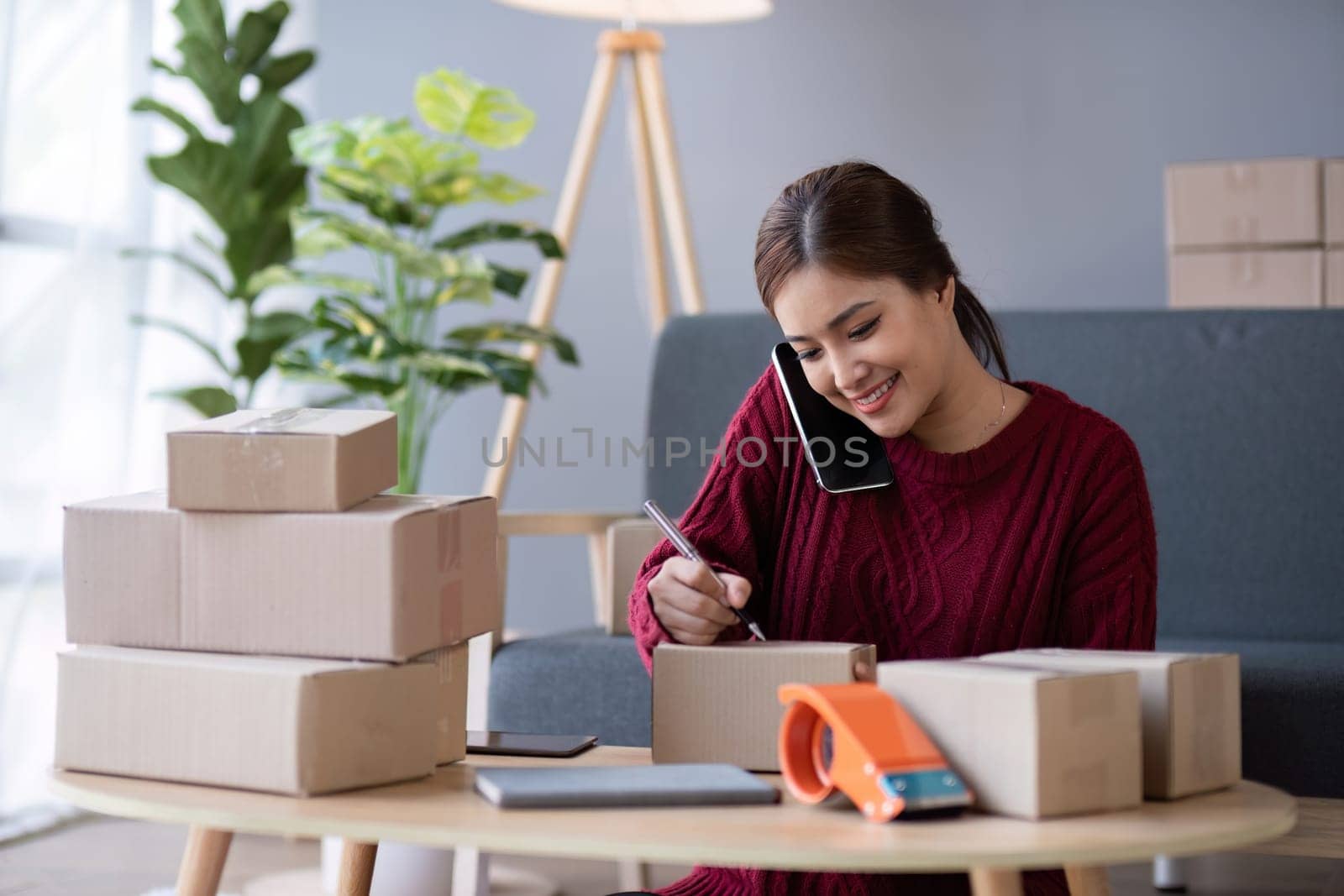 A small SME business owner receives product orders and writes shipping information on cardboard boxes in the home office to prepare for delivery..