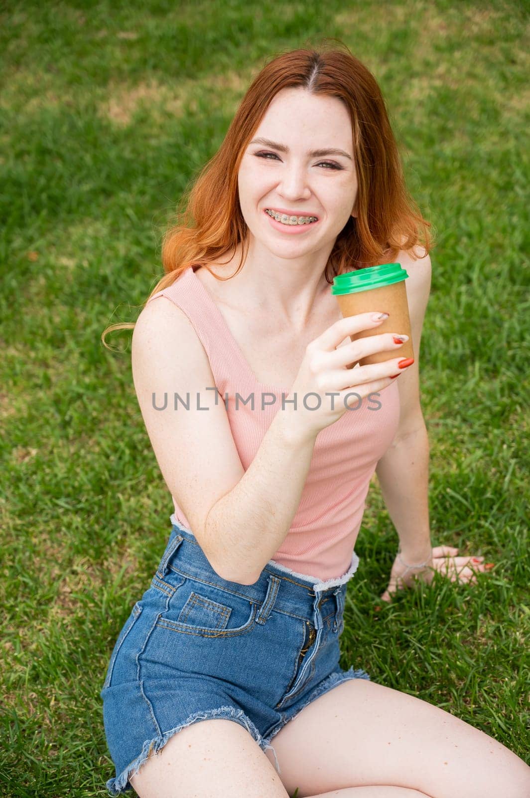 A beautiful young woman with braces on her teeth drinks from a cardboard cup while sitting on the grass. Vertical photo. by mrwed54