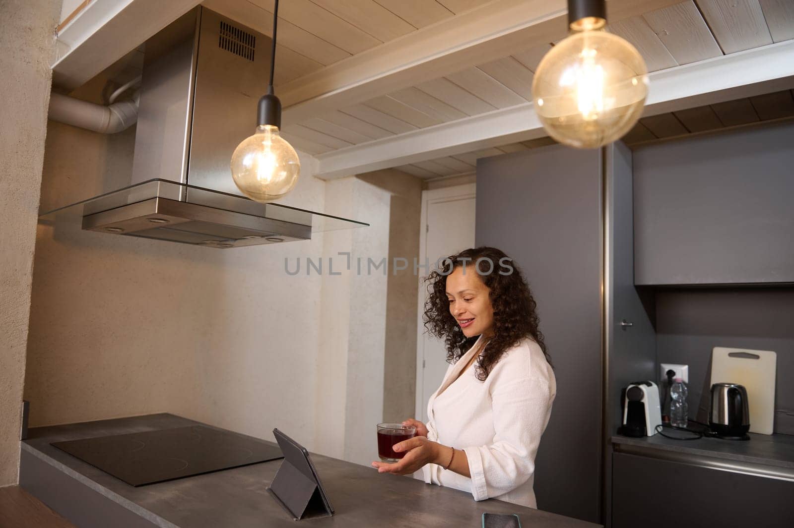 Happy pretty millennial woman in white bathrobe, relaxing at home, making a video call on digital tablet, standing at kitchen counter in modern home interior. Internet and mobile communication concept