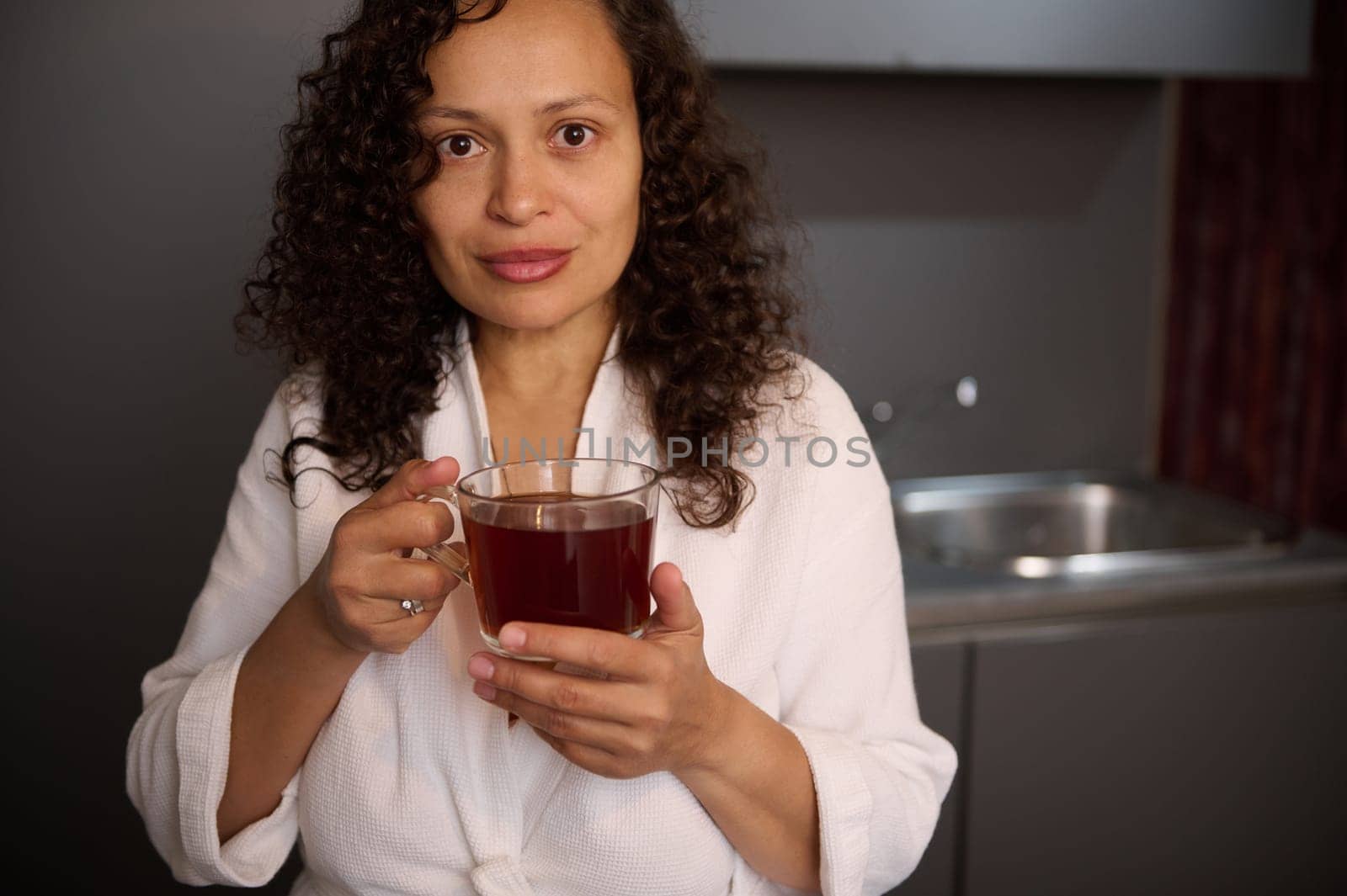 Attractive curly haired brunette in white bathrobe, holding a cup of hot tea drink, smiling looking confidently at camera. Pretty woman with mug of beverage at home interior in the morning