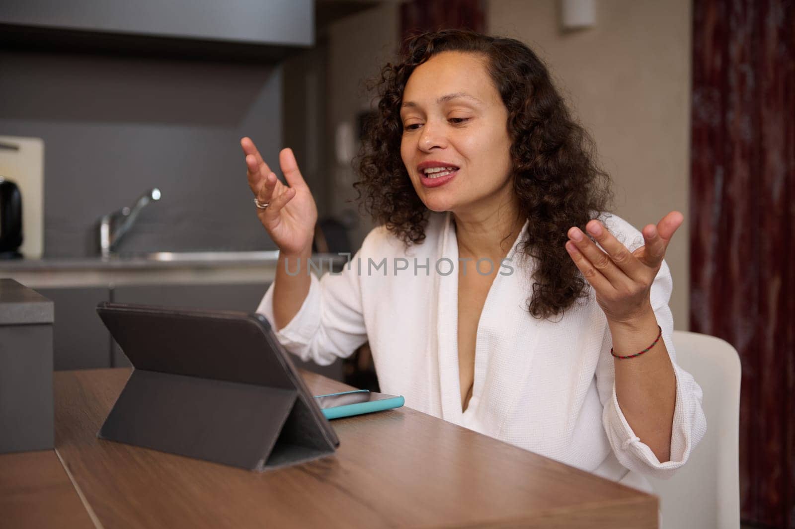 Multiracial brunette pretty woman dressed in white bathrobe, sitting at table in modern kitchen and having video call over digital tablet. People, internet, wireless technology, online communication