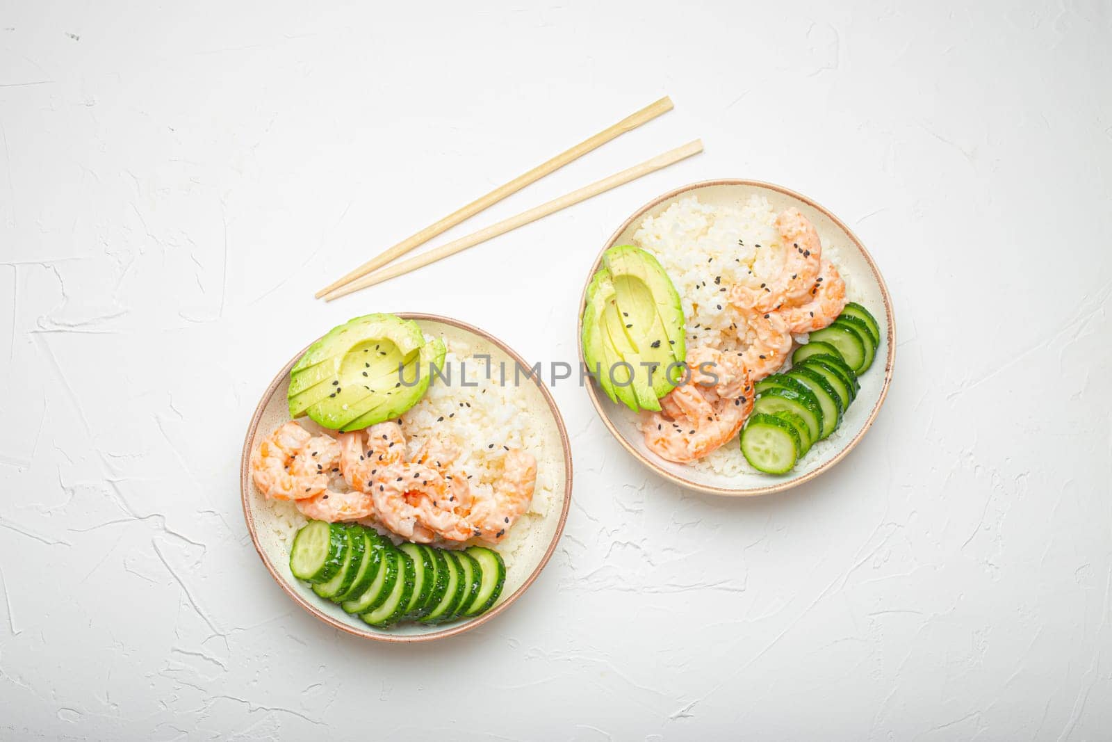 Two white ceramic bowls with rice, shrimps, avocado, vegetables and sesame seeds and wooden sticks on white concrete stone background top view. Healthy asian style poke bowl by its_al_dente